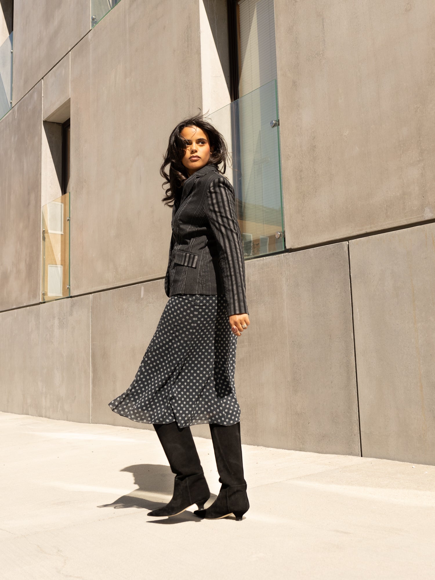Woman in a black coat and polka dot dress standing in front of a modern concrete building.