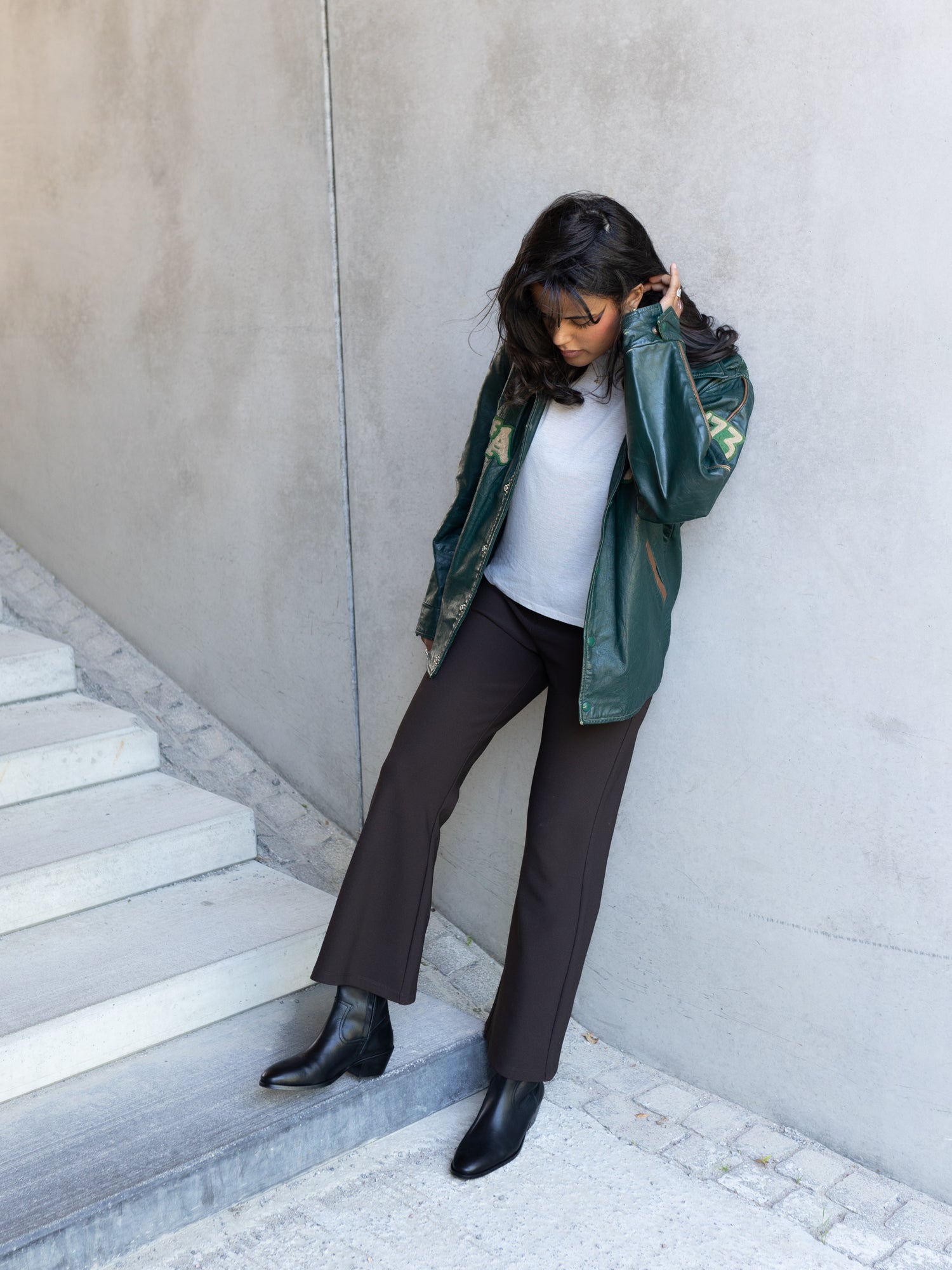 A woman leans against a concrete wall on outdoor stairs, wearing a dark green camouflage bomber jacket over a white top, dark brown wide-leg pants, and black ankle boots.