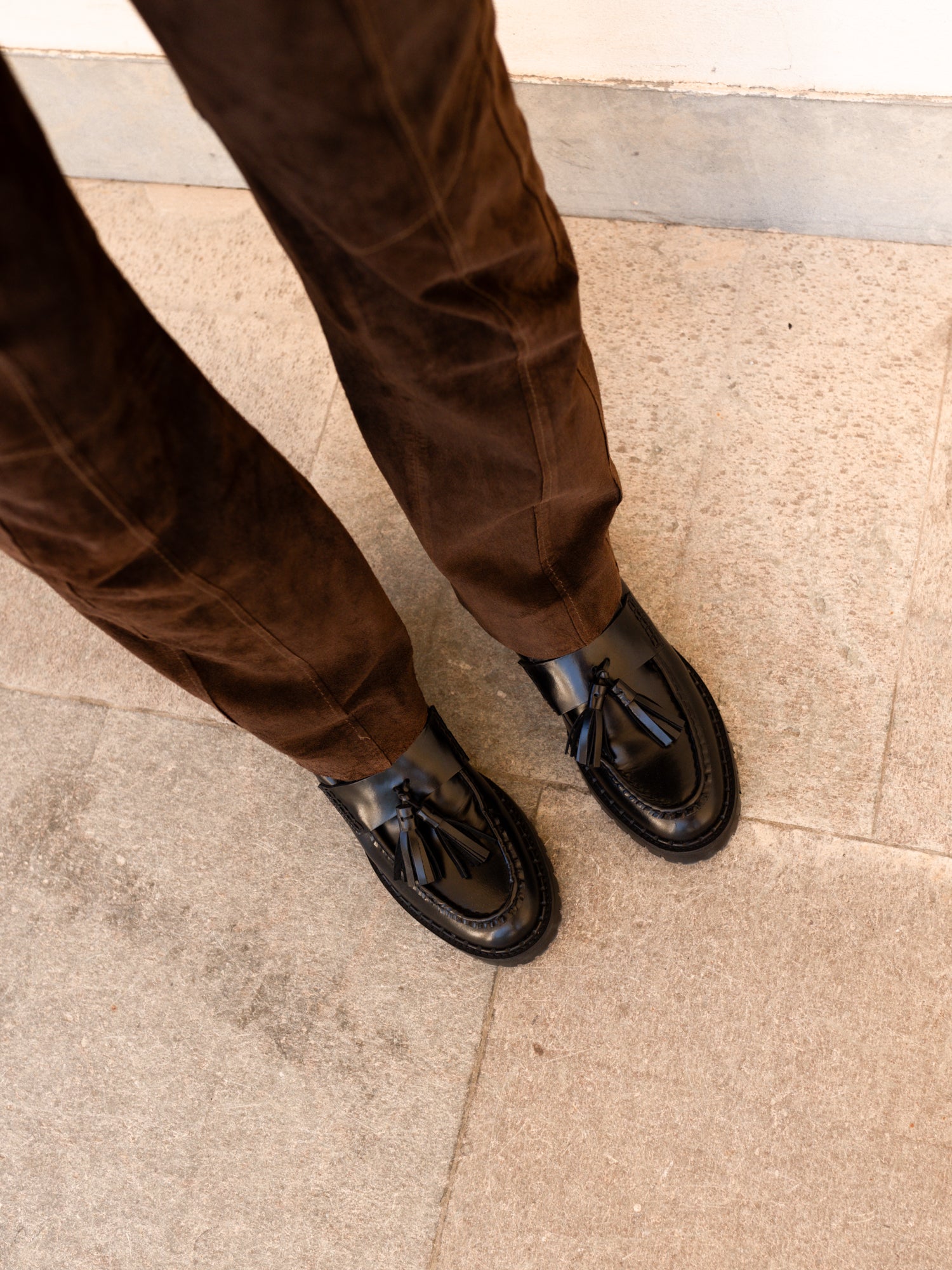 A close-up, high-angle view of a person's legs wearing brown suede trousers and black leather tassel loafers on a light beige tiled floor.