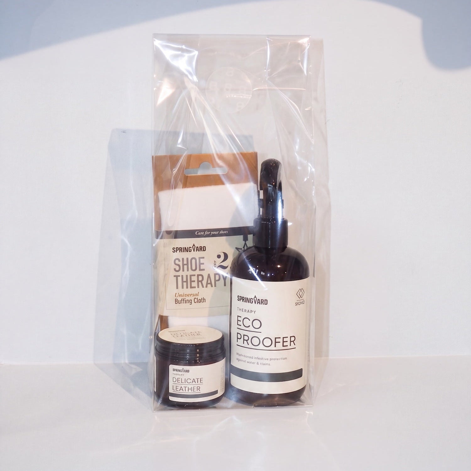 A clear plastic gift bag containing a small jar of leather cleaner, a pump bottle labeled Eco Proofer, and a box labeled Shoe Therapy, on a white surface with a light blue-gray background.