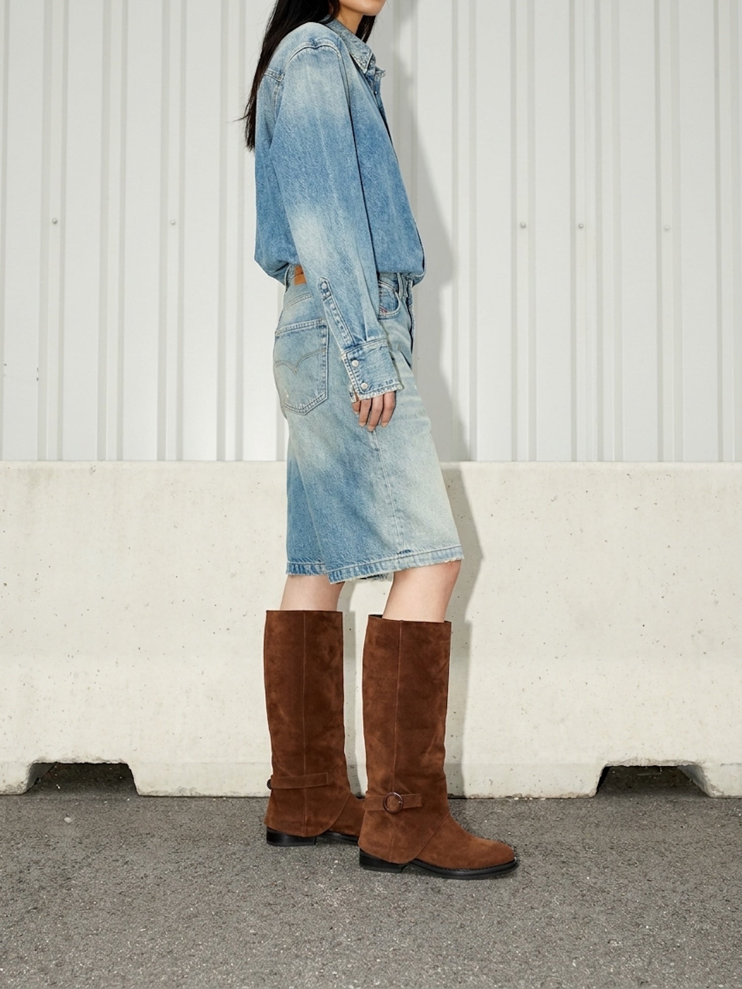 Person wearing a denim outfit with brown knee-high boots against a white wall.